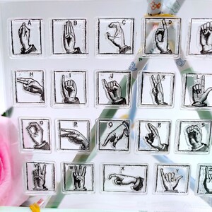 May include: A clear sheet of transparent stamps with black outlines of hands forming the letters of the alphabet in American Sign Language. The letters A through Z are represented in individual squares.