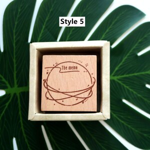 K&ouml;nnte beinhalten: Ein Stempel aus Holz mit einem kreisf&ouml;rmigen Design, das einen Planeten mit Sternen und dem Text "The memo" zeigt. Der Stempel befindet sich in einer kleinen Holzkiste.