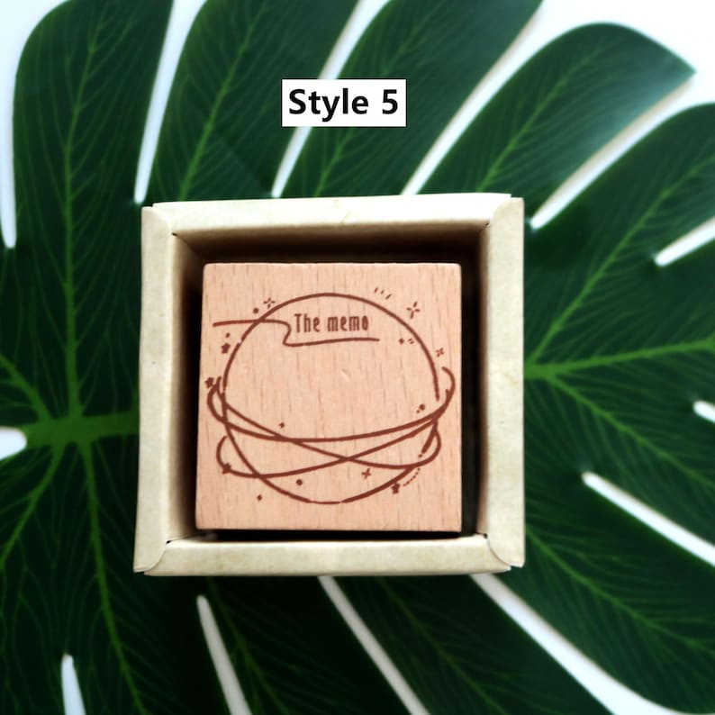 K&ouml;nnte beinhalten: Ein Stempel aus Holz mit einem kreisf&ouml;rmigen Design, das einen Planeten mit Sternen und dem Text "The memo" zeigt. Der Stempel befindet sich in einer kleinen Holzkiste.