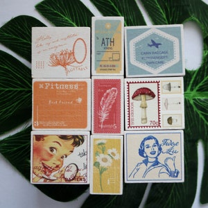 May include: Nine wooden blocks with various designs, including a mushroom, a feather, a daisy, a woman's face, a plane, and text that says "Fitness", "ATH Athens", "Cabin Baggage", "Vegetable", and "Fidere Au".