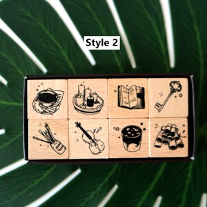 May include: A set of eight wooden stamps with black ink illustrations. The stamps feature a teacup, candles, an open book, a key, paintbrushes, a violin, a drink, and rolled items. The text "Style 2" is in the upper left corner.