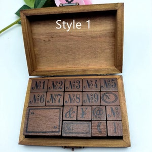 Puede incluir: Una caja de madera con tapa abatible, abierta para revelar un juego de 18 sellos de madera. Los sellos est&aacute;n dispuestos en filas y presentan varios dise&ntilde;os, incluidos n&uacute;meros, s&iacute;mbolos y un rect&aacute;ngulo en blanco. La caja est&aacute; etiquetada como "Style 1" en la parte superior.
