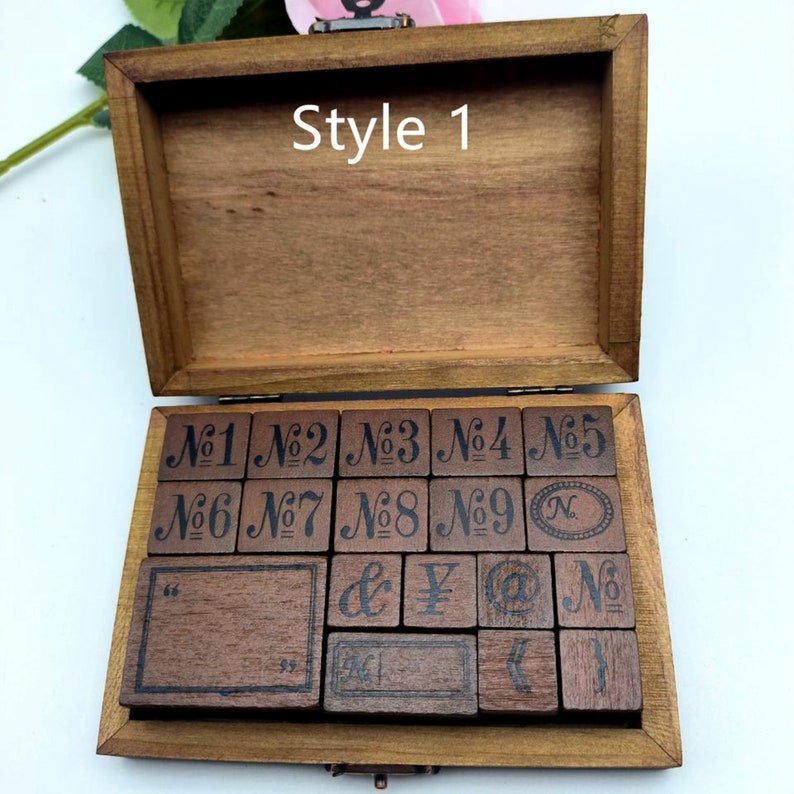 Puede incluir: Una caja de madera con tapa abatible, abierta para revelar un juego de 18 sellos de madera. Los sellos est&aacute;n dispuestos en filas y presentan varios dise&ntilde;os, incluidos n&uacute;meros, s&iacute;mbolos y un rect&aacute;ngulo en blanco. La caja est&aacute; etiquetada como "Style 1" en la parte superior.