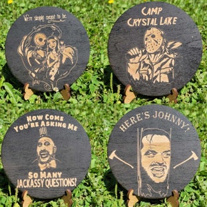 May include: Four round wooden coasters with black backgrounds and gold text and images. The coasters feature characters from popular horror movies. The first coaster features Jack Skellington and Sally from The Nightmare Before Christmas. The second coaster features Jason Voorhees from Friday the 13th. The third coaster features Captain Spaulding from House of 1000 Corpses. The fourth coaster features Jack Torrance from The Shining.