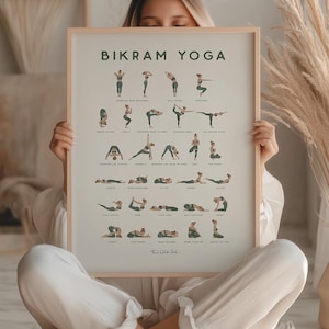 May include: A framed poster displaying "Bikram Yoga" poses, held by a person in a cross-legged position. The poster has a light wooden frame. The image features a neutral colour palette, with pampas grass and a vase in the background.