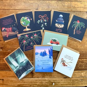 Könnte beinhalten: Eine Sammlung von Weihnachtskarten mit verschiedenen Designs. Die Karten zeigen Illustrationen einer Katze mit Hut, Palmen mit Weihnachtsmützen, Skifahrern und Winterlandschaften. Einige Karten haben Texte wie "Merry Christmas" und "Let it Snow".
