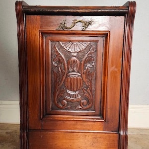May include: A wooden cabinet with a carved front panel featuring a detailed floral and urn design. The cabinet has a brass handle and is made of dark-stained wood. The sides have vertical grooves, and the cabinet sits on small feet.