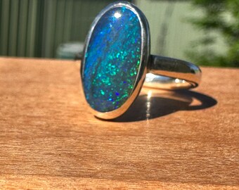 May include: A silver ring with an oval-shaped opal gemstone. The opal displays a vibrant play of color, with shades of blue and green. The ring is set on a wooden surface, with sunlight casting a shadow.