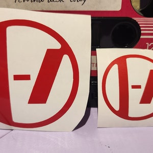 Puede incluir: Dos pegatinas rojas y blancas con un logotipo estilizado que consta de un círculo, una línea vertical, una línea horizontal y una línea diagonal. Las pegatinas son de diferentes tamaños y están sobre una superficie blanca.