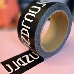 Może przedstawiać: Rolka czarnej taśmy washi z białym, pikselowanym tekstem. Taśma jest częściowo rozwinięta, odsłaniając mały kawałek taśmy z tym samym tekstem. Taśma leży na różowej powierzchni.