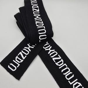 May include: A black fabric strap featuring a repeating white pixelated text pattern that reads "TOUJZUXOZDJOU". The strap is folded into a layered, overlapping arrangement, showcasing the woven texture of the material and the high-contrast white lettering.