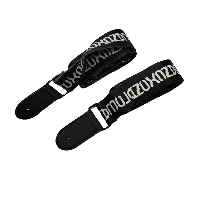 K&ouml;nnte beinhalten: Zwei schwarze Gitarrengurte mit wei&szlig;em "ZUXDZ"-Text. Sie haben schwarze Lederenden und Metallbeschl&auml;ge. Die Gurte sind nach oben links und unten rechts abgewinkelt.