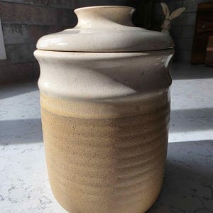 May include: A cylindrical ceramic canister with a lid, featuring a two-tone glaze. The upper portion and lid are a smooth, glossy off-white with fine crackle details, while the lower body is a textured, matte tan with horizontal ridges.
