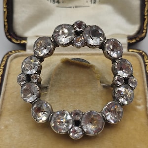 May include: A silver brooch with a circular design of clear faceted stones. The brooch is in a brown velvet box.