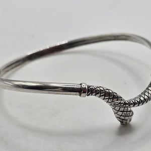 Peut inclure: Un bracelet jonc argenté avec un motif de serpent. Le bracelet présente une bande lisse et incurvée qui se termine par une tête de serpent avec des écailles texturées. La tête de serpent est positionnée à l'extrémité du bracelet.