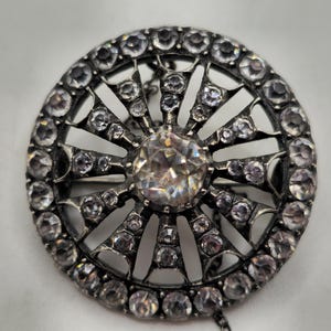 May include: A circular, silver-coloured brooch featuring a large, clear central gemstone. The brooch is surrounded by smaller, clear gemstones and has an intricate, openwork design. The brooch is vintage in style.