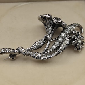 Op de afbeelding: Een zilverkleurige metalen broche in de vorm van een bloemenboeket, voorzien van meerdere stelen en bloemhoofdjes ingelegd met talloze heldere, rondgeslepen strass-steentjes. Het metaal heeft een donkere, geoxideerde afwerking in de groeven.