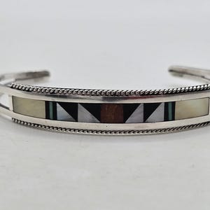Op de afbeelding: Een zilveren manchetarmband met een ingelegd ontwerp. De armband heeft een centraal rechthoekig paneel met geometrische patronen in zwart, bruin, wit en turquoise. De randen zijn versierd met een gedraaid touwontwerp.