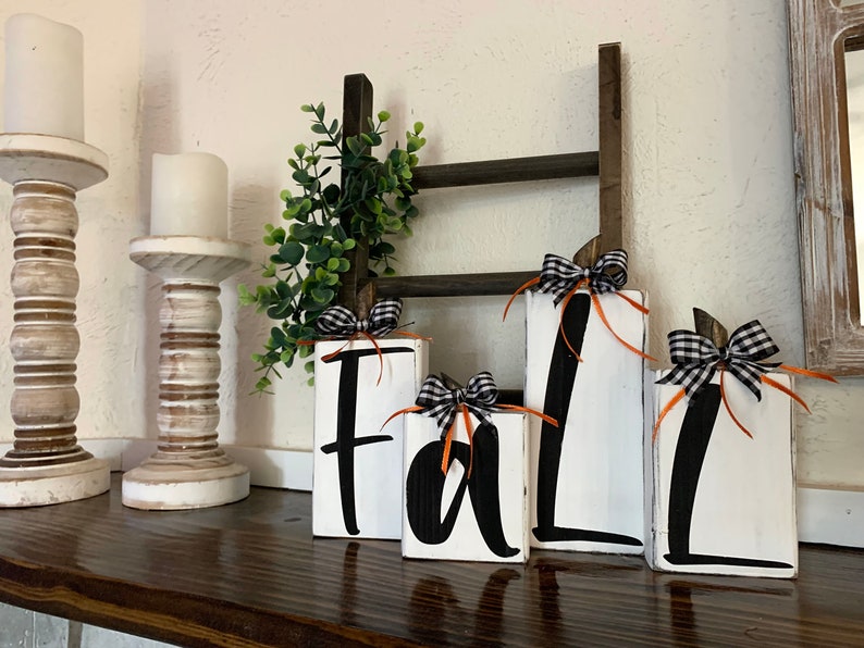 May include: Four white wooden blocks with black lettering spelling out the word 'fall.' Each block is decorated with a black and white checkered ribbon and an orange ribbon. The blocks are arranged on a wooden surface.