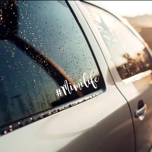 May include: A silver car with a white vinyl sticker on the back window that says "#minilife". The car is parked outside and there are raindrops on the window.