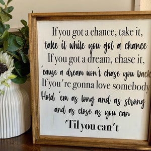 May include: A wooden sign with white paint and black lettering. The sign reads: "If you got a chance, take it, take it while you got a chance. If you got a dream, chase it, 'cause a dream won't chase you back. If you're gonna love somebody, hold 'em as long and as strong and as close as you can 'Til you can't."