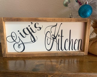 May include: A wooden sign with a white background and black lettering. The sign says "Gigi's Kitchen" in a cursive font.