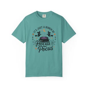 Peut inclure: Un t-shirt à manches courtes bleu sarcelle avec un motif noir représentant deux sorcières sur des balais, un chaudron avec du liquide violet, des toiles d'araignée, des étoiles et un croissant de lune. Le texte indique "IT'S JUST A BUNCH OF HOCUS POCUS".