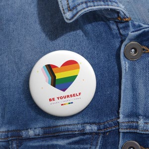 May include: A round white pin-back button featuring a heart-shaped Progress Pride flag design. Below the heart, the text reads BE YOURSELF in red, followed by the words proudly, loudly, freely in smaller black font and a small rainbow colour bar.