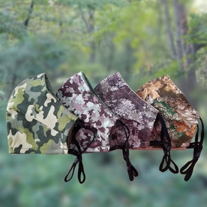 May include: Four camouflage face masks in various patterns, including green, brown, and purple, are displayed against a blurred green background. Each mask has black adjustable ear loops. The masks are designed for protection and style.