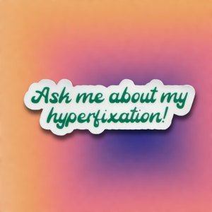 Op de afbeelding: Witte sticker met groene tekst die "Vraag me over mijn hyperfixatie!" zegt.