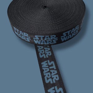 May include: A roll of black ribbon featuring the "STAR WARS" logo in blue. The ribbon is tightly wound, with a portion unrolled, displaying the repeating text. The background is a solid blue.
