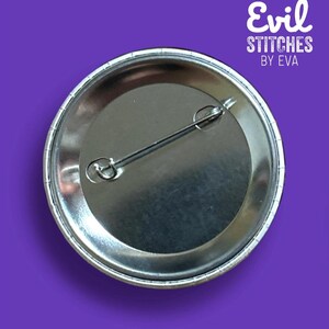 May include: The back of a silver button with a pin attached. The button has a white border and the text "Evil Stitches by Eva" is printed on the back.