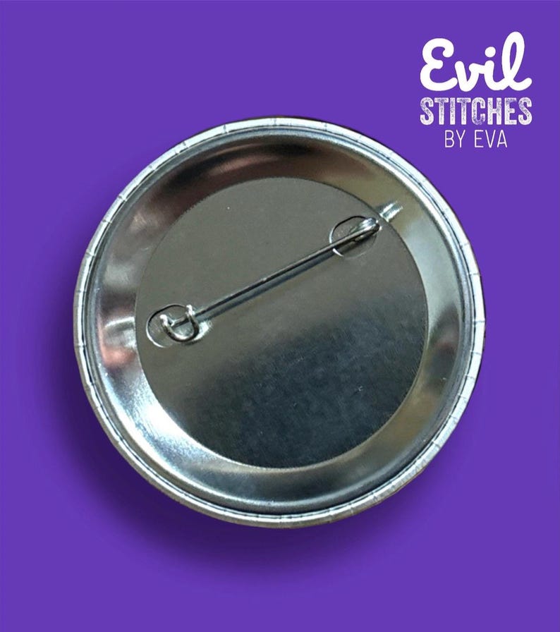 May include: The back of a silver button with a pin attached. The button has a white border and the text "Evil Stitches by Eva" is printed on the back.