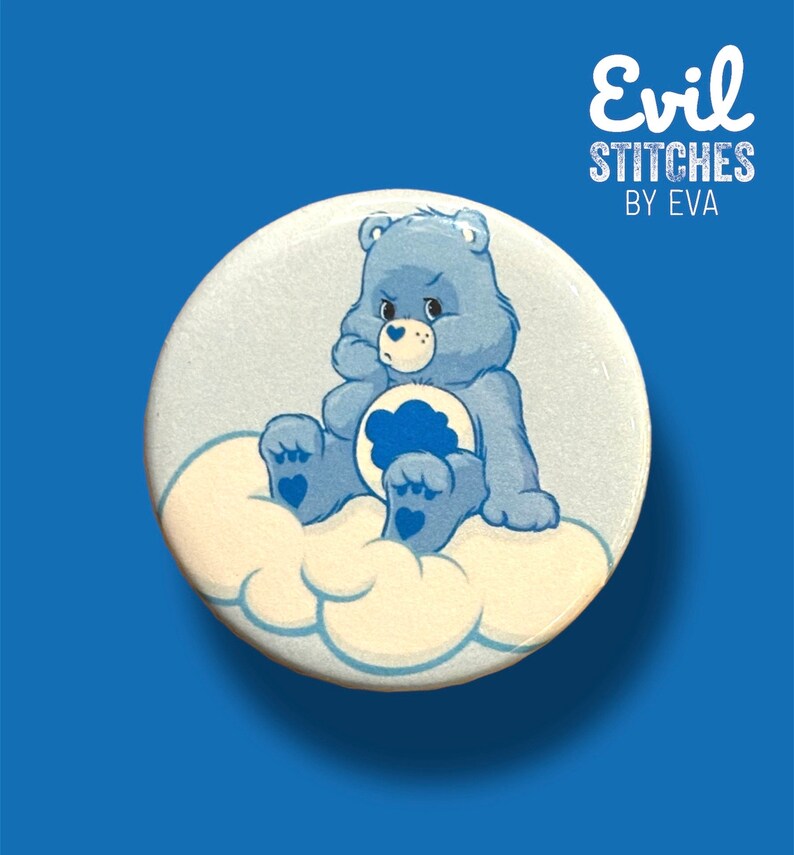 May include: A blue Care Bear with a sad expression sits on a white cloud. The bear is holding a blue cloud in its paws. The text "Evil Stitches by Eva" is written in white on a black background above the bear.