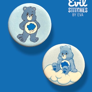 Könnte beinhalten: Zwei blaue, von Care Bear inspirierte Anstecknadeln mit einem mürrischen Ausdruck. Die erste Nadel zeigt einen blauen Bären mit einer blauen Wolke und Regentropfen auf seinem Bauch. Die zweite Nadel zeigt einen blauen Bären, der auf einer weißen Wolke sitzt. Beide Nadeln tragen den Text "Evil Stitches by Eva".