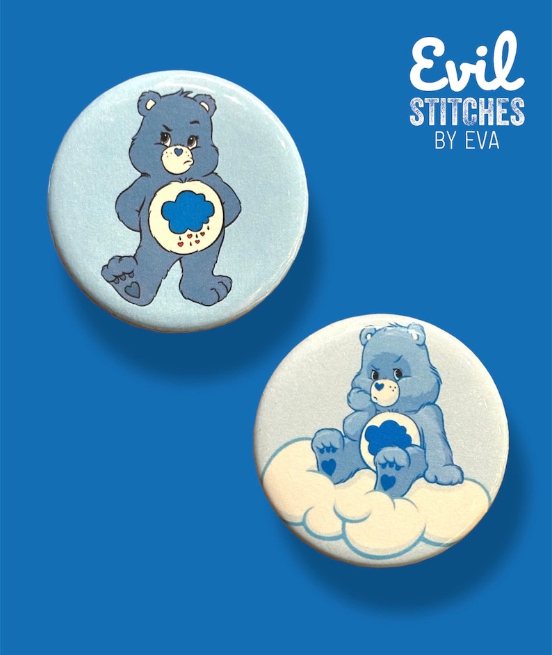 May include: Two blue Care Bear-inspired button pins with a grumpy expression. The first pin features a blue bear with a blue cloud and raindrops on its belly. The second pin features a blue bear sitting on a white cloud. Both pins have the text "Evil Stitches by Eva" on them.