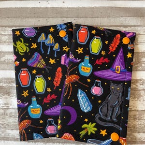 May include: Two black fabric wristlets with a witchy Halloween theme. The fabric features a black background with colorful images of potion bottles, spiders, stars, moons, mushrooms, feathers, and a black cat. Each wristlet has a single orange button.