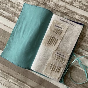 May include: An open teal fabric book with two white felt pockets. Each pocket contains a set of needles labeled "Tapestry 18" and "Tapestry 20".