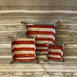May include: Three decorative fabric baskets with a patriotic American flag design. The baskets are red and white striped with blue star accents on the corners. The baskets are different sizes and have small handles.