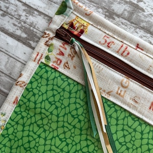 May include: Close-up of a fabric pouch with a brown zipper and decorative ribbons. The pouch features a green pebble pattern and a cream-colored fabric with printed text and illustrations. The edges are trimmed with a floral pattern.