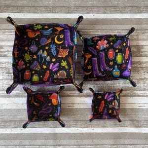 May include: Four black fabric dice trays with a witchy theme. The trays are lined with a black fabric with a colorful print of various witchy items, including boots, potions, mushrooms, and stars. The trays are in different sizes and are arranged in a square shape.