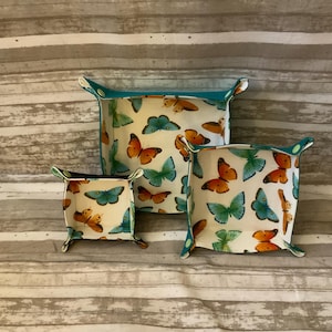 May include: Set of three fabric catchall trays with a butterfly pattern. The trays are white with orange, teal, and green butterflies. The edges are teal. The trays are different sizes, with the largest measuring approximately 8 inches square.