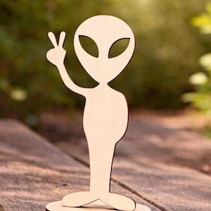 Op de afbeelding: Een houten alienfiguur met een vredesteken. De lichtgekleurde alien heeft grote ovale ogen en is gemonteerd op een ronde, donkerbruine basis. De figuur is gemaakt van lichtgekleurd hout en staat op een houten oppervlak.