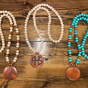 May include: Three wooden bead necklaces with large wooden pendant charms. The necklaces are in white, turquoise, and brown beads. The charms are engraved with the letters "AHS".