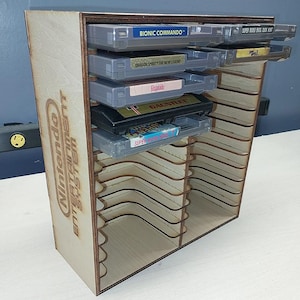 May include: A light-colored wooden storage rack for video game cartridges featuring multiple horizontal slots. The side panel is laser-engraved with the text Nintendo Entertainment System. Several gray and black game cartridges are inserted.