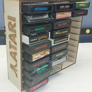 May include: A light-colored wooden storage rack with the word ATARI laser-cut into the side, holding multiple black rectangular video game cartridges. The cartridges feature various game titles including Asteroids, Pitfall, and Pac-Man.