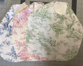 May include: Three baby bibs featuring a white background with a repeating toile pattern. One bib has a blue print, one has a red print, and one has a green print. Each bib displays detailed illustrations of children playing outdoors in nature.