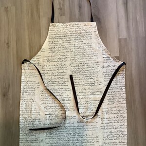 以下が含まれることがあります： 黒い紐の付いた白いエプロン。エプロンは黒い手書きの文字のパターンで覆われています。