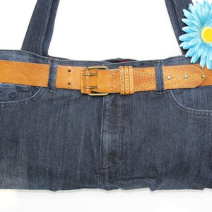 Könnte beinhalten: Eine Denim-Tragetasche mit einem braunen Ledergürtel mit Metallnieten. Die Tasche hat eine blaue Blume oben.