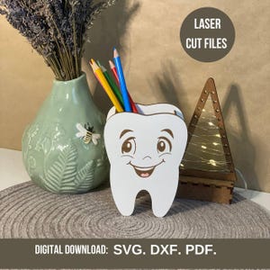 May include: A white, tooth-shaped pencil holder with a smiling face, holding colorful pencils. A green vase with a bee design and a wooden Christmas tree with lights are also in the image. Text reads "LASER CUT FILES" and "DIGITAL DOWNLOAD: SVG. DXF. PDF."